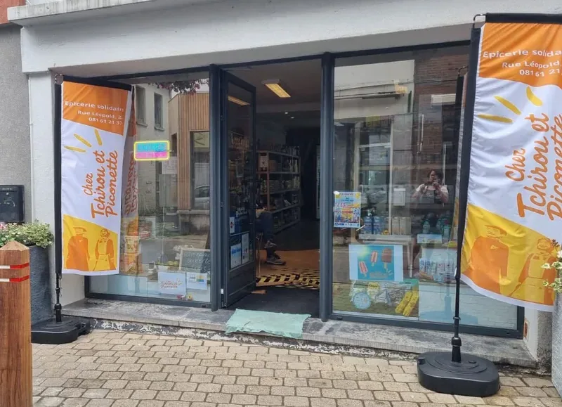 Deux drapeaux publicitaires orange et blanc encadrant l'entrée d'une épicerie solidaire.